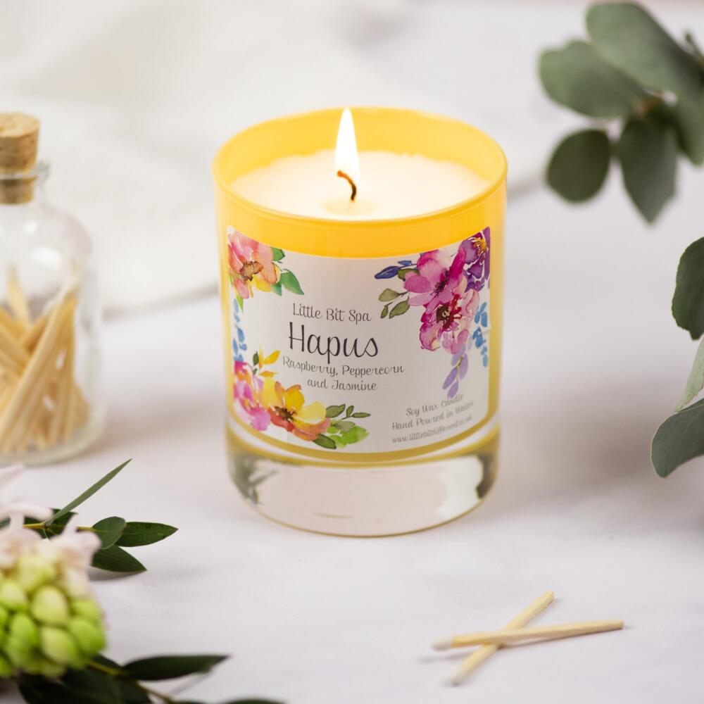 yellow glass candle with welsh language label with flowers in the background from the Little Bit Spa range.  The fragrance is raspberry, peppercorn and Jasmine